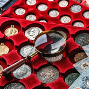A collection of antique coins in red velvet packaging. A magnifying glass is sitting on top of the collection.