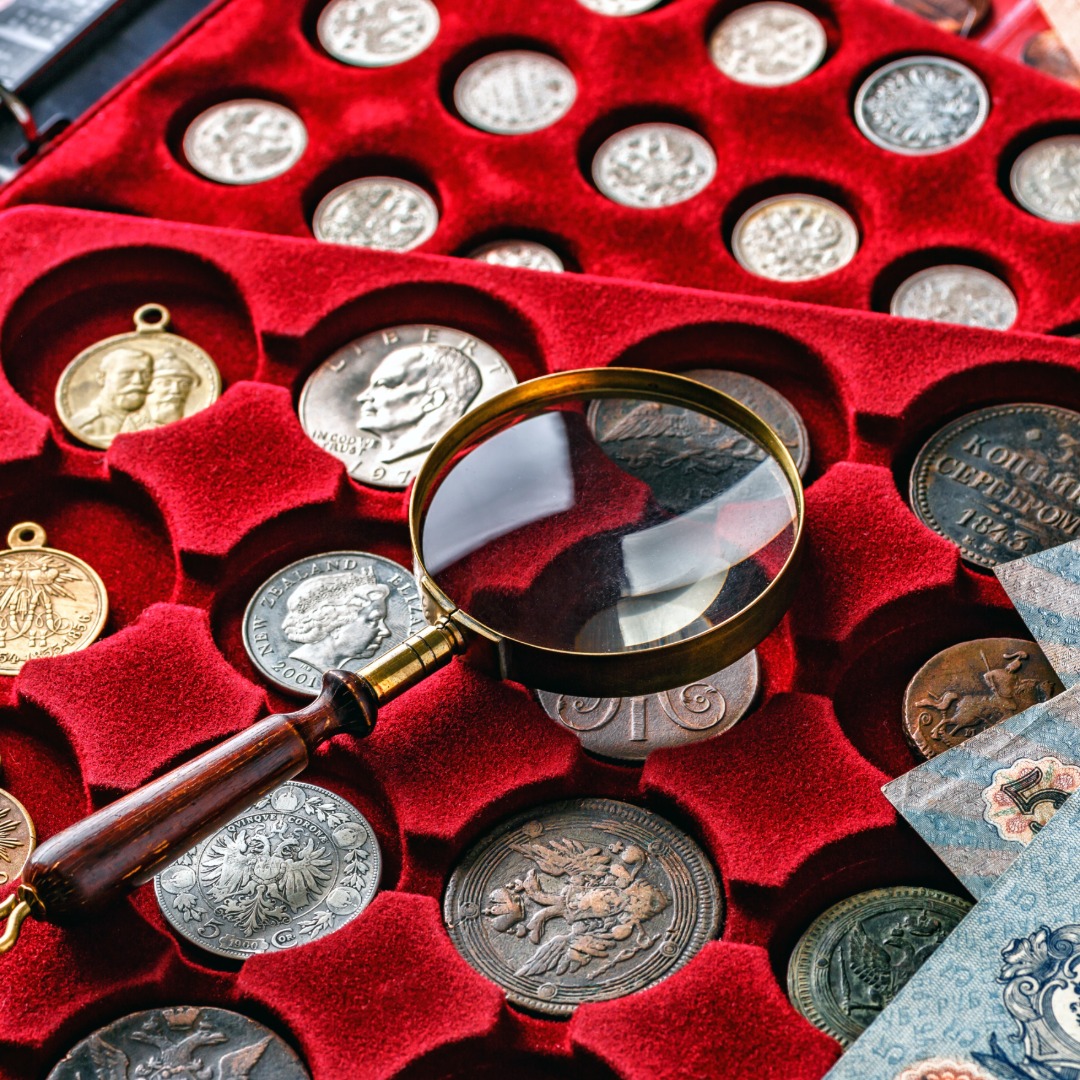 A collection of antique coins in red velvet packaging. A magnifying glass is sitting on top of the collection.