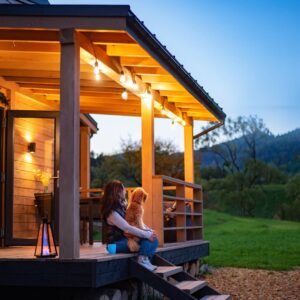 A woman sits on a wooden cabin porch holding a dog under string lights, overlooking a grassy field and mountains at dusk.