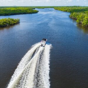 A small motorboat leaves a white wake on a wide river bordered by green mangroves under a cloudy sky.