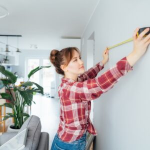 A young woman holding an extended tape measure up to a blank white wall behind a couch in her home.