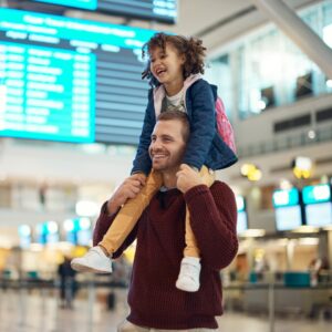 A man stands in an airport holding a child on his shoulders while both of them smile and stand near flight displays.