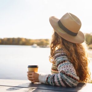 A woman in a hat sits by a lakeside, holding a coffee cup and looking out over calm water on a sunny day.