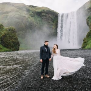 A man in a tux and a woman in a wedding dress hold hands and stand on the bank of a river with a waterfall behind them.
