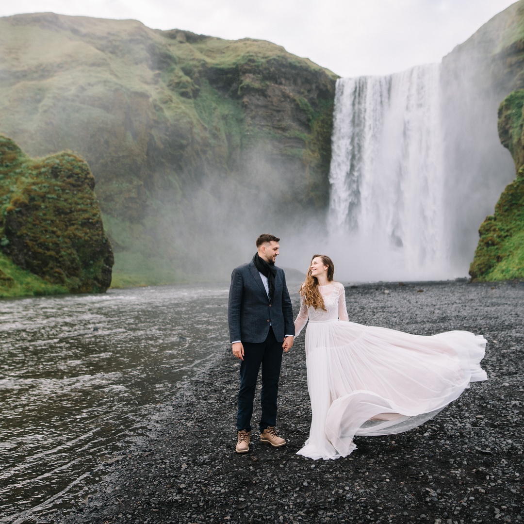 A man in a tux and a woman in a wedding dress hold hands and stand on the bank of a river with a waterfall behind them.