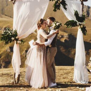 A couple embracing beneath white drapes and greenery while holding flowers on a sunny mountain landscape.