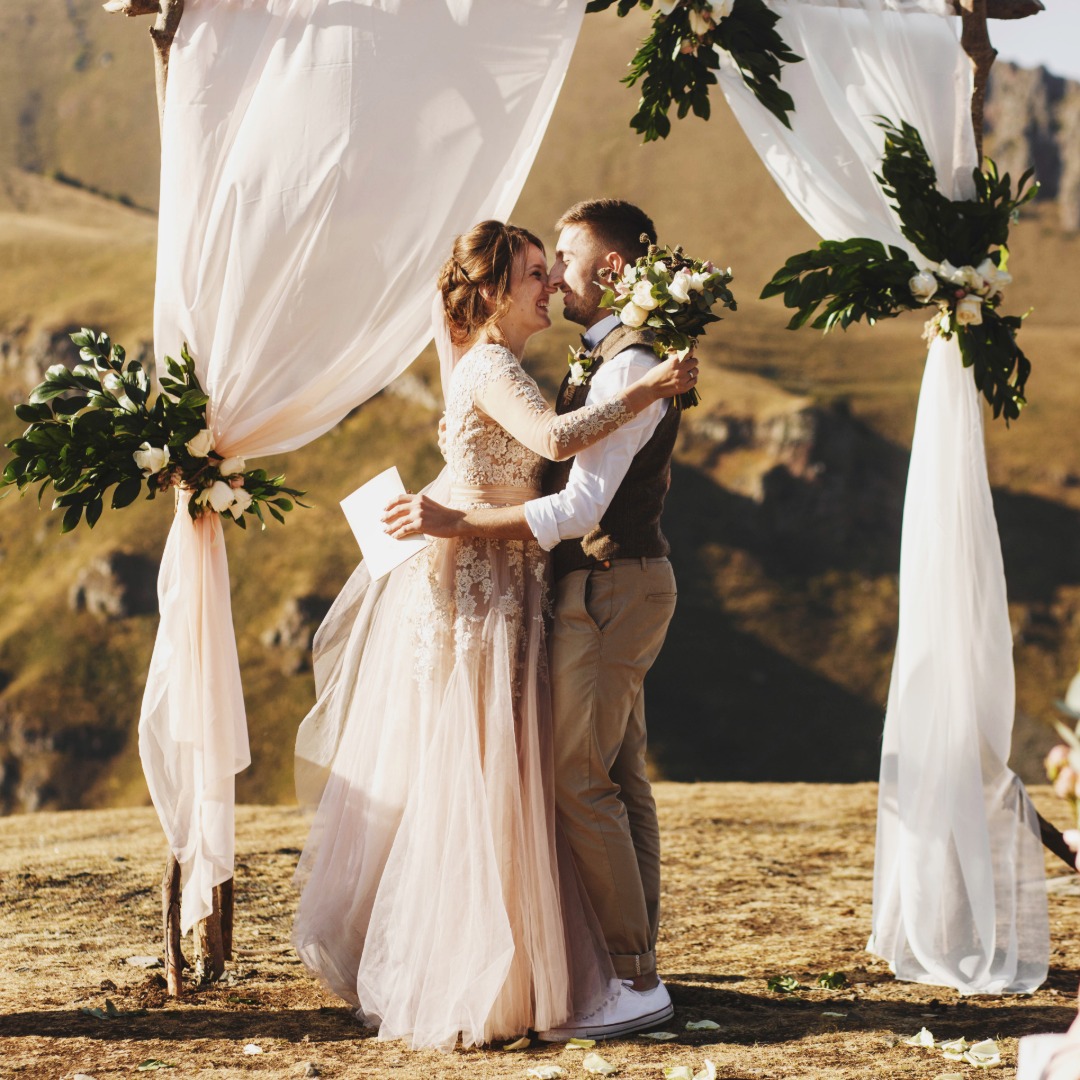 A couple embracing beneath white drapes and greenery while holding flowers on a sunny mountain landscape.