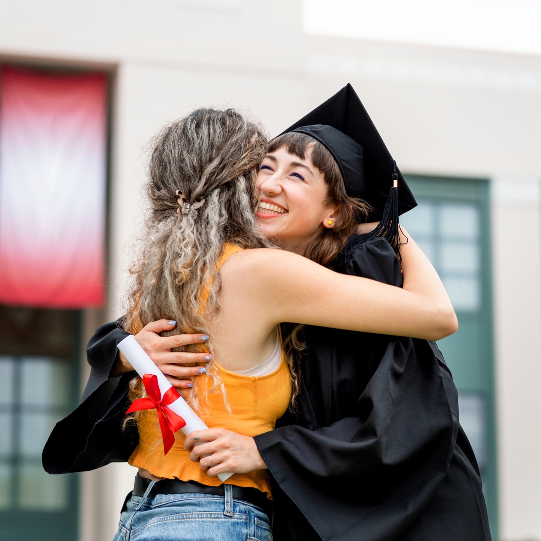 A smiling graduate in a cap and gown hugging another person outdoors during a graduation ceremony with trees behind them.