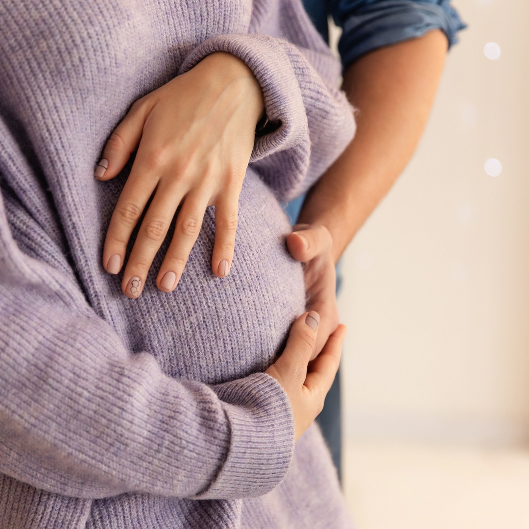 A pregnant woman is holding her belly while a man behind her is reaching around to also place a hand on her belly.