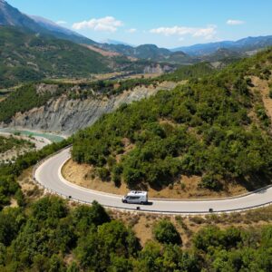 An aerial view of a van driving on a curvy road through a mountainous landscape filled with tons of trees.