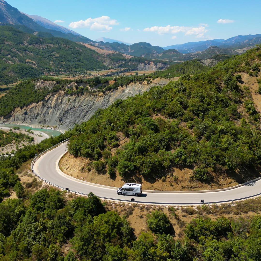 An aerial view of a van driving on a curvy road through a mountainous landscape filled with tons of trees.