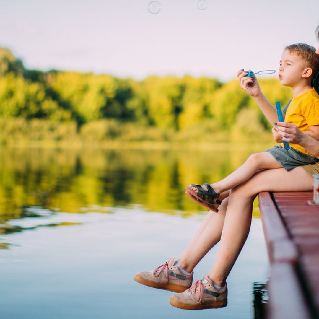 A woman and young child sitting on a wooden dock by the lake while blowing bubbles with a blue bubble wand.
