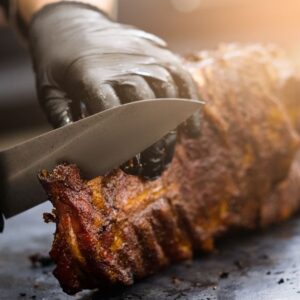 A close view of black-gloved hands slicing cooked ribs with a knife on a metal surface under warm light.