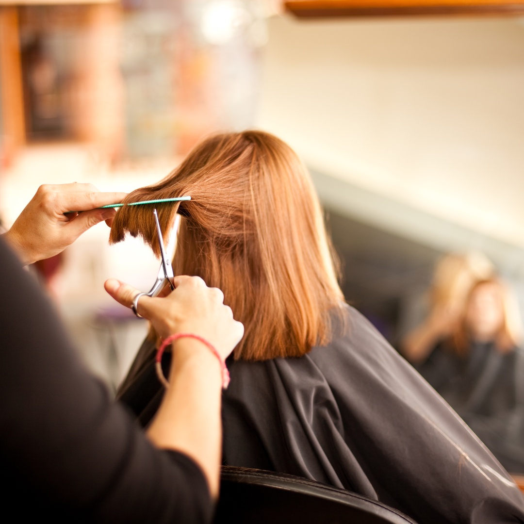 A hairdresser trims a client's straight red hair with scissors and a comb in a salon as the client sits before a mirror.