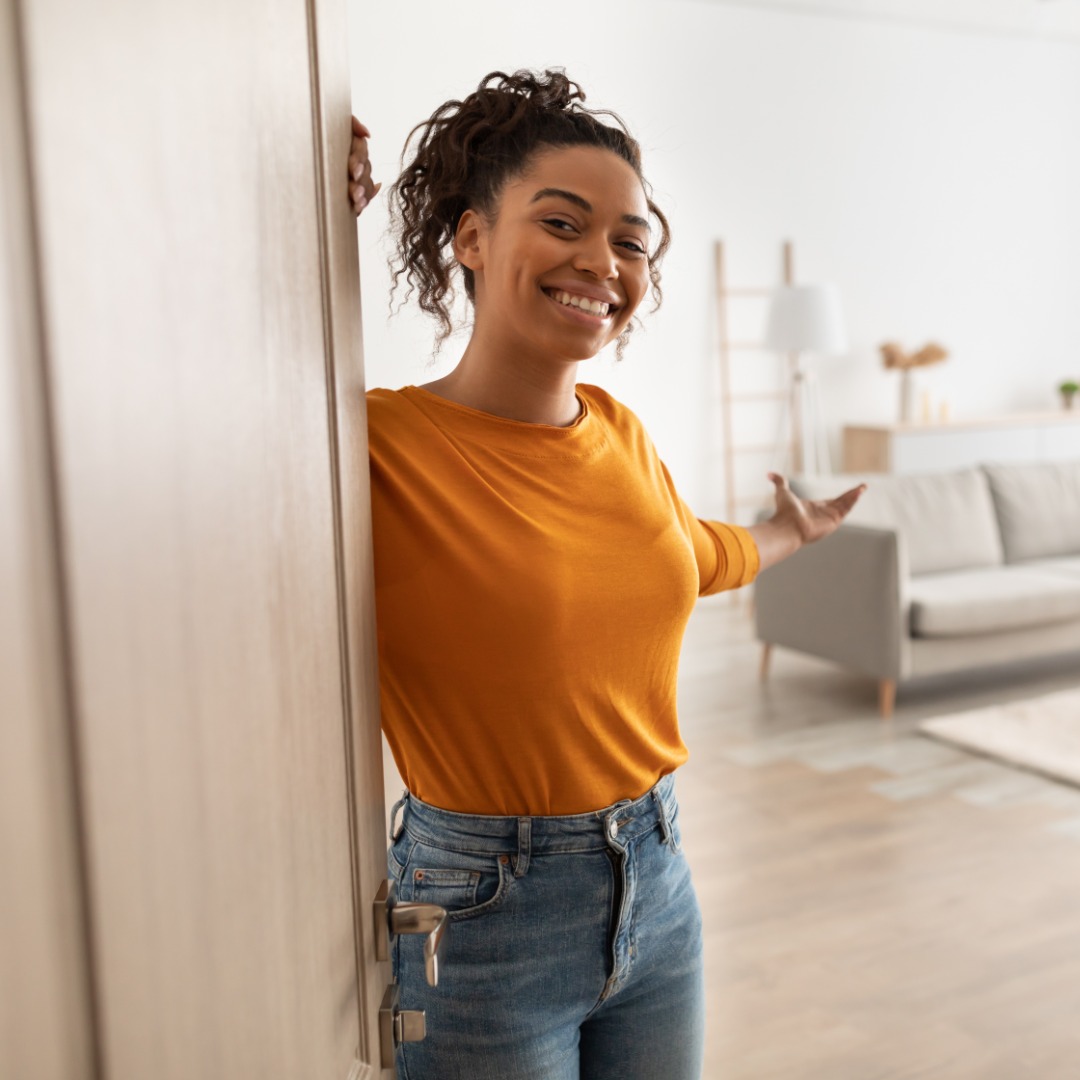 A cheerful woman opening the door to her home with her arm spread out to welcome the guest into the open space.