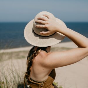 A woman wearing a sunhat and brown top, holding the hat while standing near beach grass with water in the background.