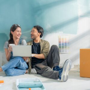 A happy couple sits on the floor against a semi-painted wall in their home. They laugh together with design plans.