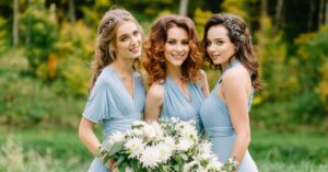 Three women wearing light blue formal dresses smile together outdoors, holding white floral bouquets in a garden setting.
