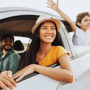 A woman is in the driver’s seat of a car. There is a man in the passenger’s seat and a child sticking their hands out the window.