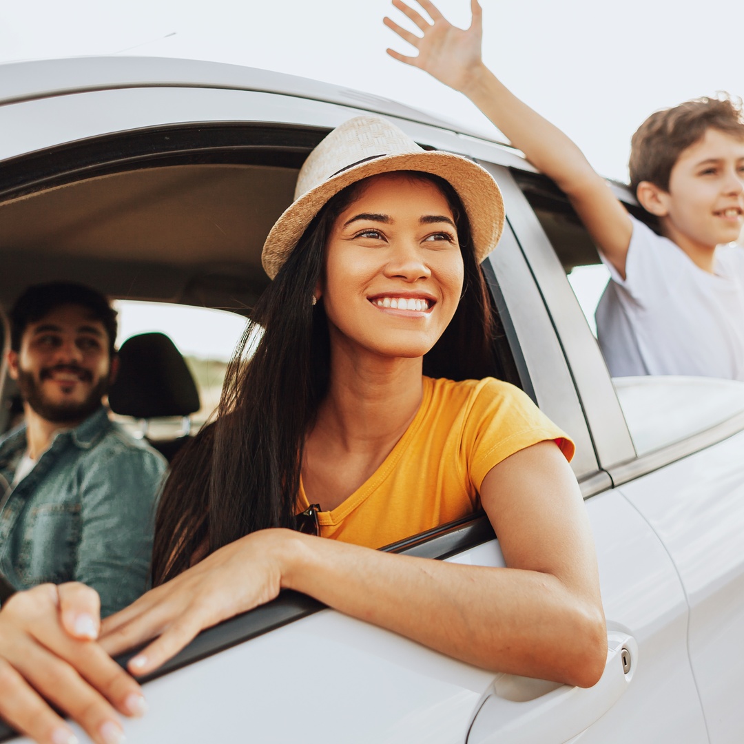 A woman is in the driver’s seat of a car. There is a man in the passenger’s seat and a child sticking their hands out the window.