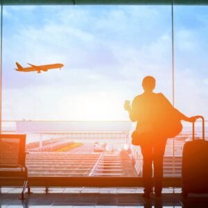 A woman with a suitcase and a coffee in her hand is standing next to a large window as she watches an airplane take off.