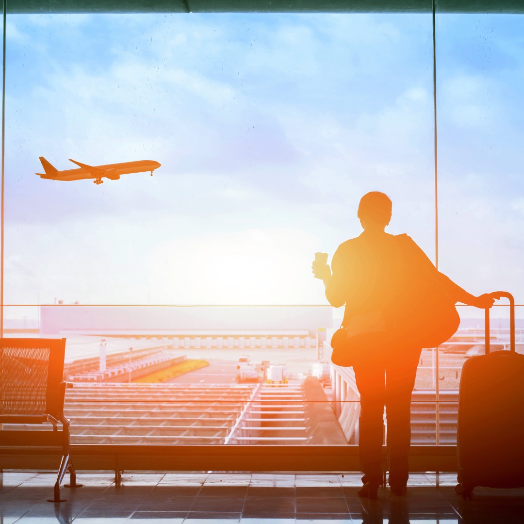 A woman with a suitcase and a coffee in her hand is standing next to a large window as she watches an airplane take off.