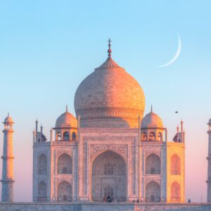 A sunset view of the Taj Mahal with a tiny sliver of a moon behind it in the sky. The sunset lights the building orange.