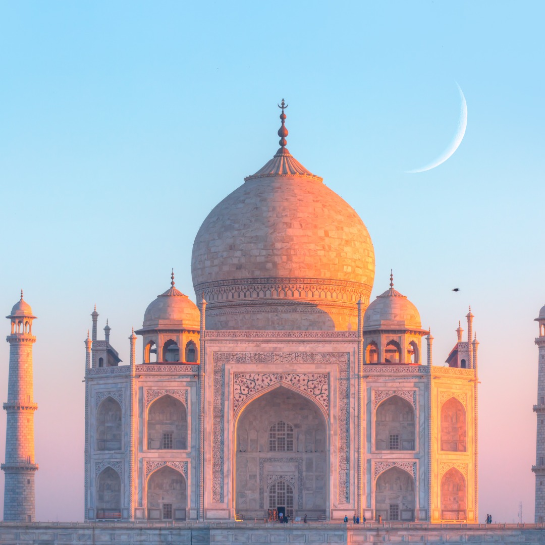 A sunset view of the Taj Mahal with a tiny sliver of a moon behind it in the sky. The sunset lights the building orange.