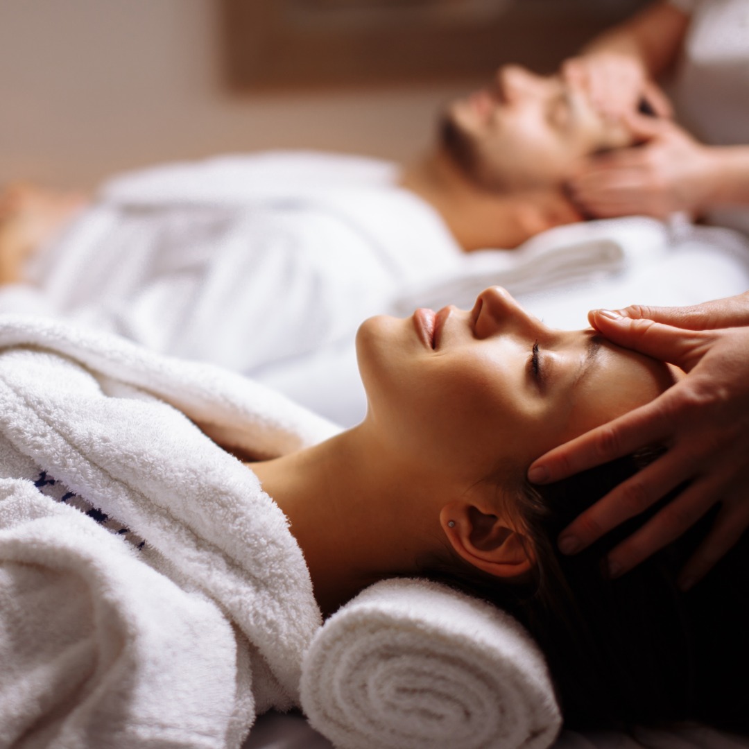 Two people relax in spa robes while therapists provide calming head massages in a peaceful spa setting.