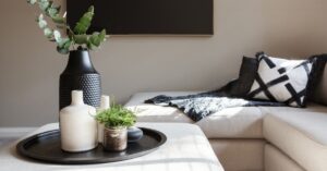 A close-up of a living room coffee table with a sectional sofa behind. There's a large black vase with greenery on the table.