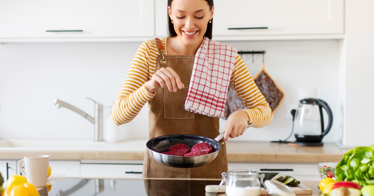 A woman wearing a brown apron stands in the kitchen, sprinkling salt into a pan that contains two uncooked steaks.
