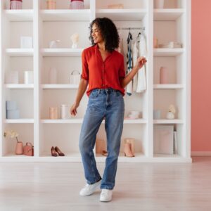 A woman in a red top, blue jeans, and white sneakers stands beside a mirror and wardrobe while choosing an outfit.