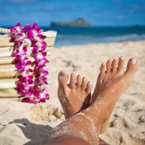 Bare feet rest in the sand beside a striped beach bag draped with a purple orchid lei, with the blue ocean nearby.