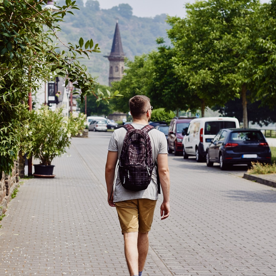 A traveler with a backpack walks along a quiet street lined with trees, homes, parked cars, and mountain views.
