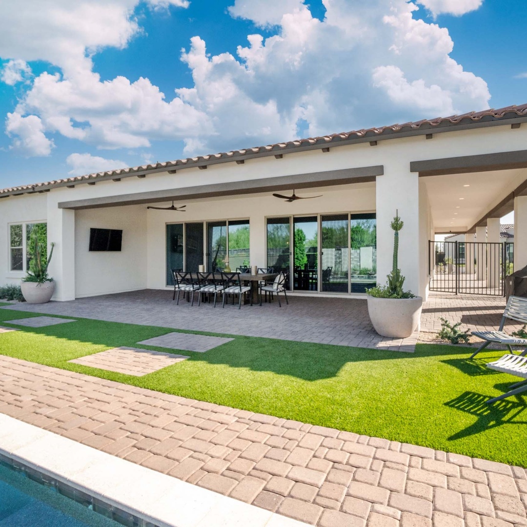 A spacious covered patio beside a swimming pool, with a dining table, lounge chairs, and a green lawn outside a white house.