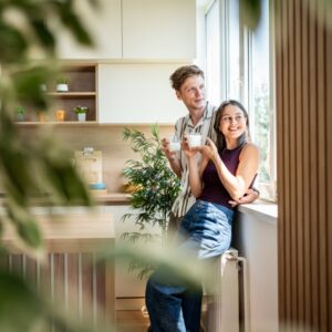 A man in a striped shirt and a woman in a sleeveless purple top holding white mugs beside a sunlit kitchen window.