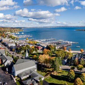Aerial view of a lakeside town with marinas, waterfront buildings, colorful autumn trees, and a blue lake under clouds.