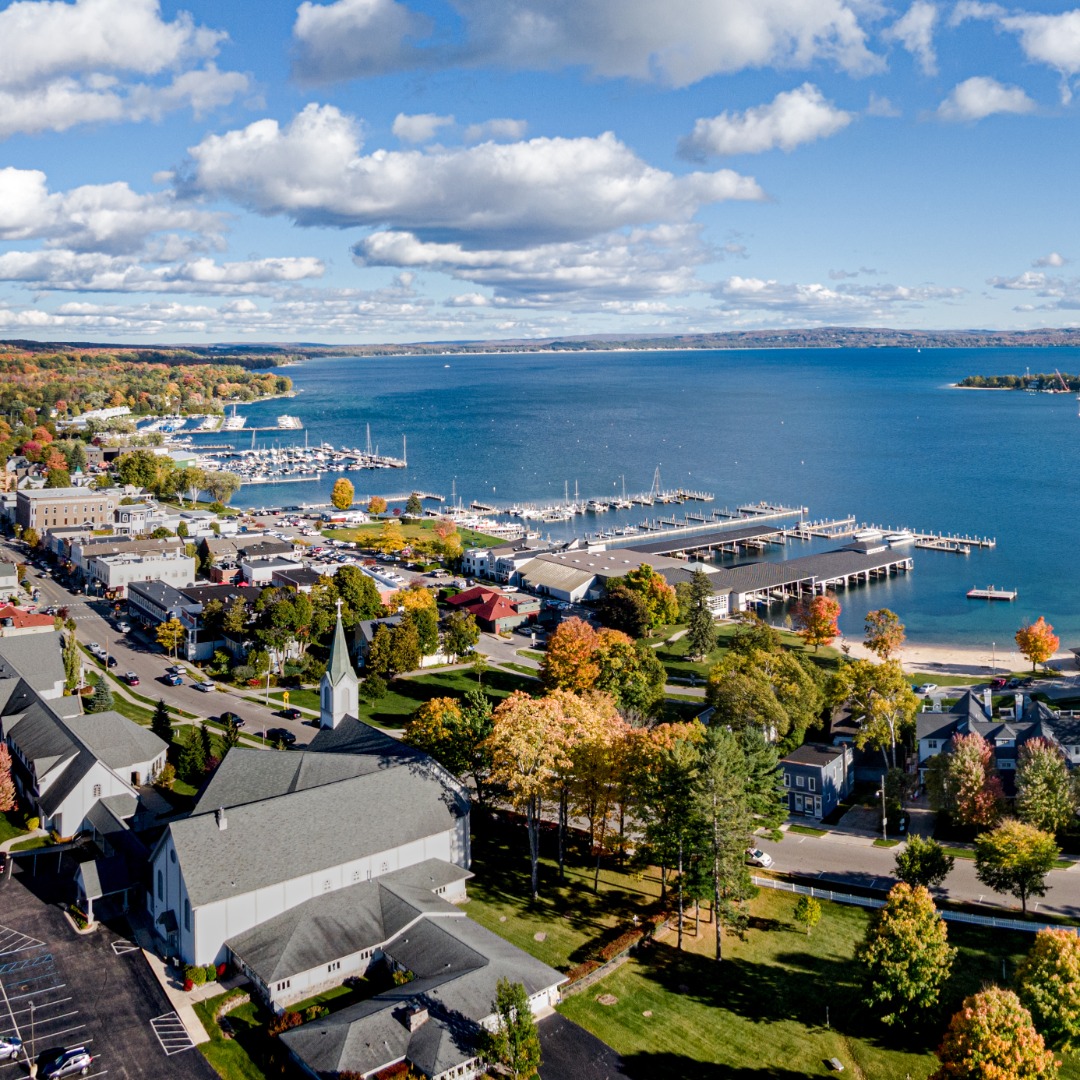 Aerial view of a lakeside town with marinas, waterfront buildings, colorful autumn trees, and a blue lake under clouds.